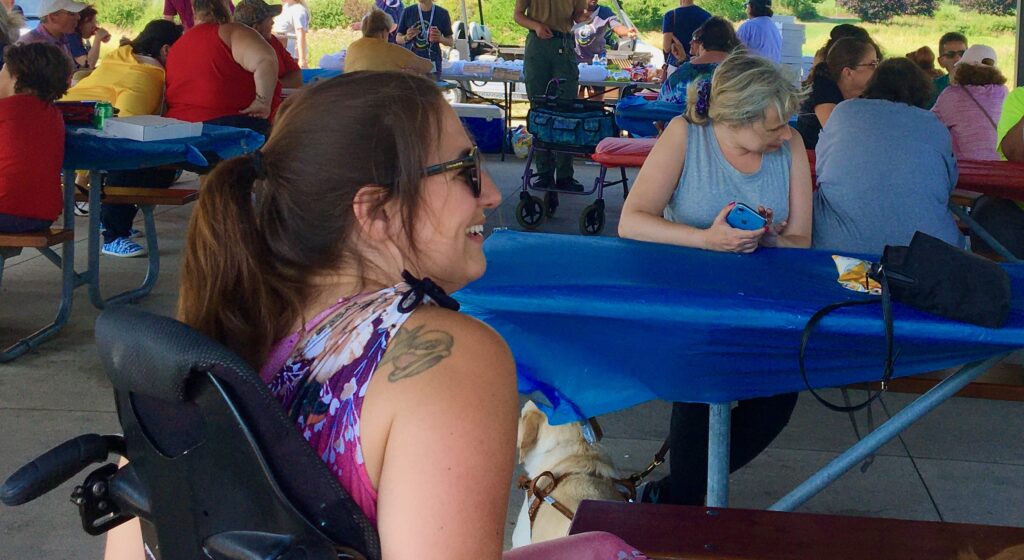 Image of woman sitting in a wheelchair smiling facing a crowd of guests at the park pavilion